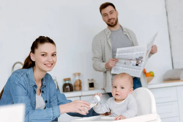 Madre alimentación hijo con leche en biberón en la cocina, padre con periódico detrás - foto de stock