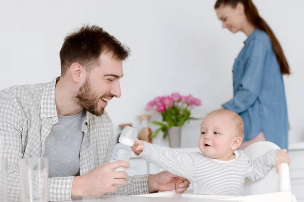 Улыбающийся отец кормит младенца молоком в детской бутылочке, мать стоит на кухне позади — стоковое фото