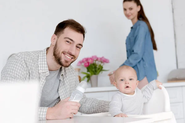 Padre alimentación hijo con leche en biberón, madre de pie en la cocina detrás - foto de stock