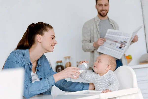 Mãe feliz alimentando filho com leite em garrafa de bebê na cozinha, pai com jornal atrás — Fotografia de Stock