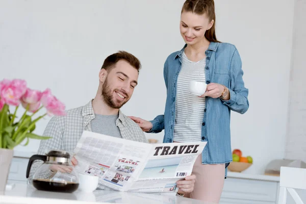 Mann liest Reisezeitung, während lächelnde Frau mit Kaffee in der Küche steht — Stockfoto