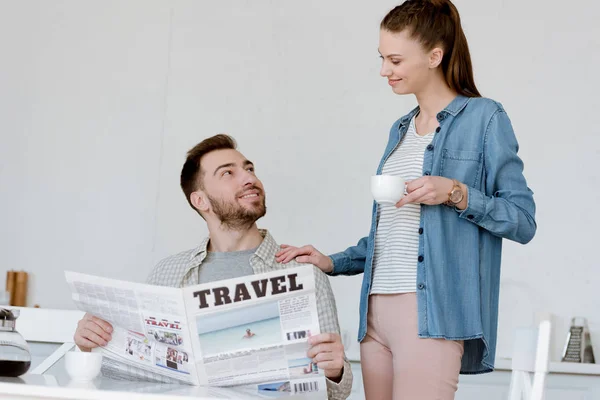 Mann liest Reisezeitung, während Frau mit Kaffee in der Nähe steht — Stockfoto