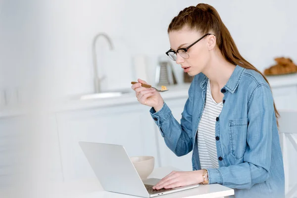 Young female freelancer using laptop on kitchen while eating breakfast — Stock Photo