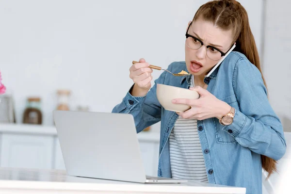 Shocked female teleworker eating breakfast while talking on smartphone and looking on laptop — Stock Photo