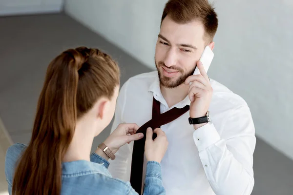 Esposa usando una corbata en el marido mientras él habla en el teléfono inteligente - foto de stock