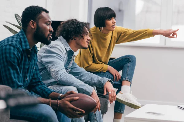 Young mixed race woman pointing by finger to multiethnic male friends and watching american football match — Stock Photo