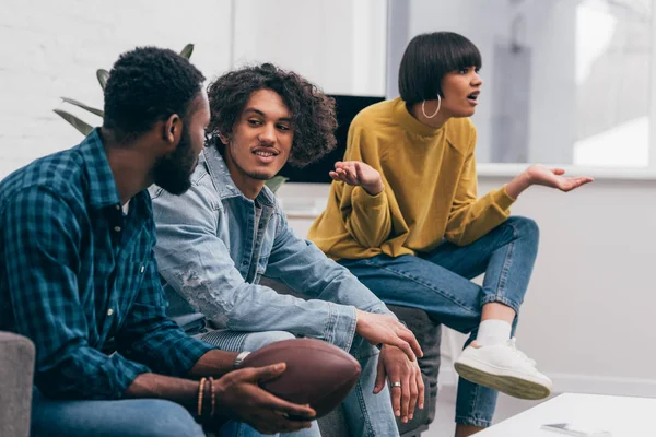 Amis masculins multiculturels parlant les uns aux autres et choqué femme de race mixte avec de larges bras regarder match de football américain — Photo de stock