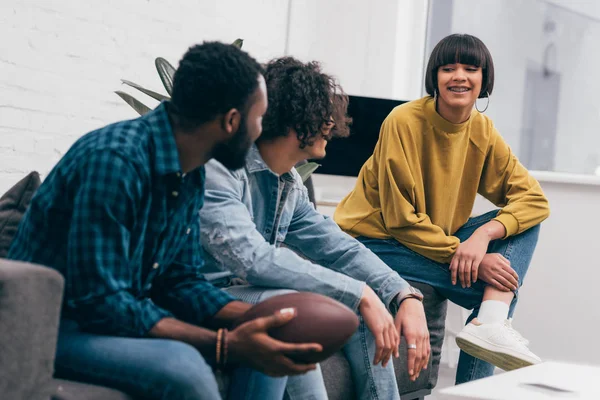 Young smiling multiethnic friends sitting on couch with ball for american football and talking to each other — Stock Photo