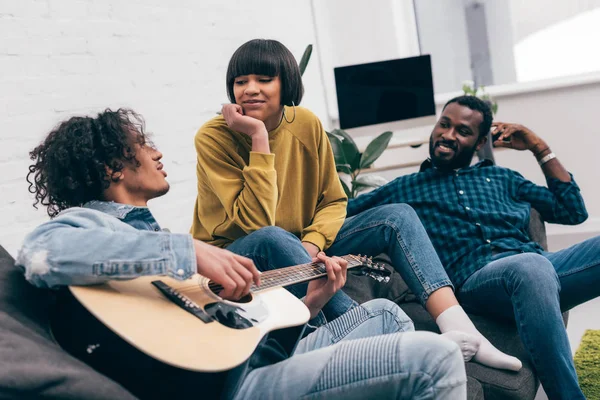 Homme de race mixte jouant à la guitare acoustique à des amis multiethniques sur le canapé — Photo de stock