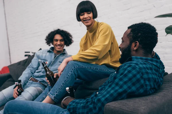 Smiling multicultural group of friends with bottles of beer talking and sitting on couch — Stock Photo