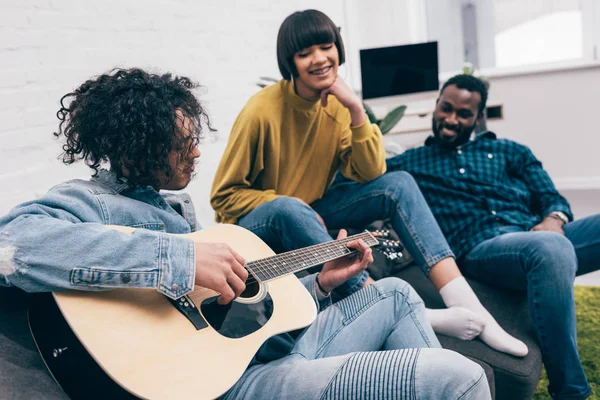 Вид сбоку на молодого человека смешанной расы, играющего на гитаре перед мультикультурными друзьями — стоковое фото