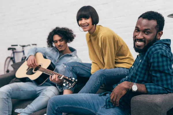 Grupo multicultural de amigos sentados en el sofá y tocando la guitarra - foto de stock