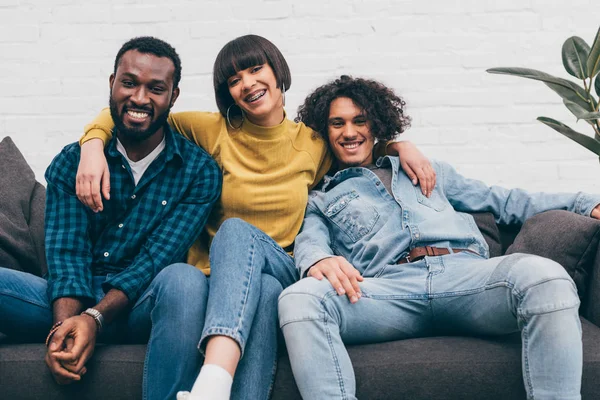 Mujer de raza mixta abrazando dos amigos masculinos sonrientes en el sofá - foto de stock