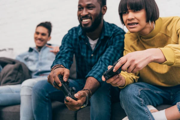 Amigos multiétnicos con joysticks jugando videojuego y joven sentado detrás - foto de stock