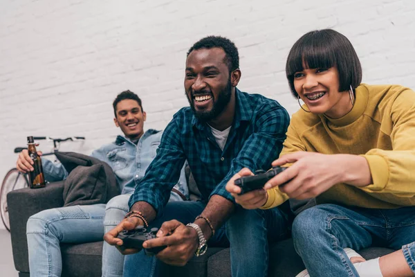 Sonriente hombre afroamericano y mujer de raza mixta con joysticks jugando videojuego y joven sentado detrás en el sofá con botella de cerveza - foto de stock