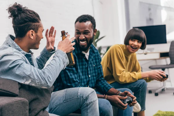 Joven hombre de raza mixta con botella de cerveza haciendo un gesto a un amigo afroamericano y una joven sentada detrás - foto de stock