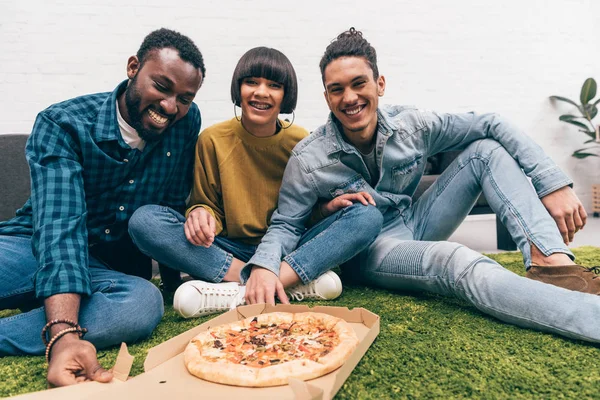 Grupo sonriente de amigos multiétnicos comiendo pizza y sentados en la alfombra - foto de stock