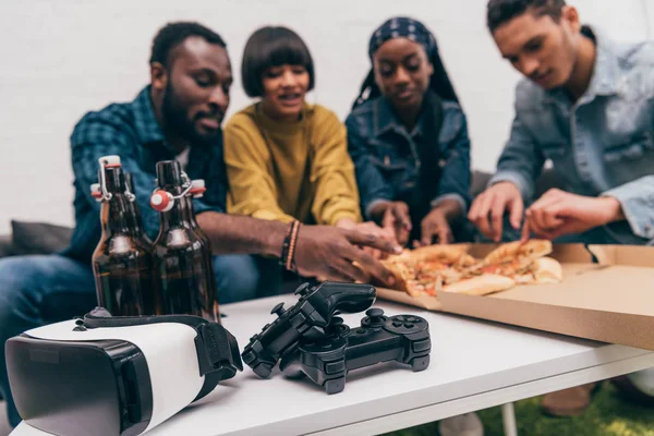 Primer plano joysticks tiro, botellas de cerveza y auriculares de realidad virtual con grupo de amigos detrás de comer pizza - foto de stock