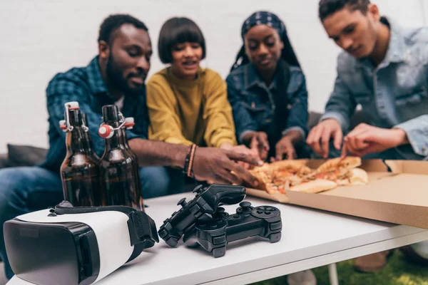 Closeup shot joysticks, bottles of beer and virtual reality headset with group of friends behind eating pizza — Stock Photo