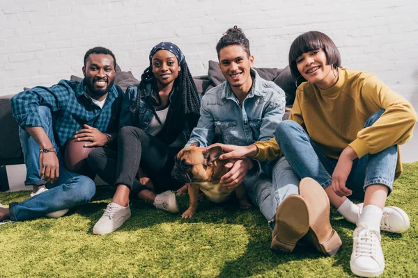 Young smiling multicultural friends sitting on rug with french bulldog — Stock Photo