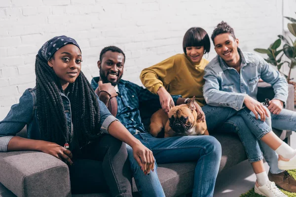 Groupe de jeunes amis multiethniques assis sur le canapé avec chien — Photo de stock