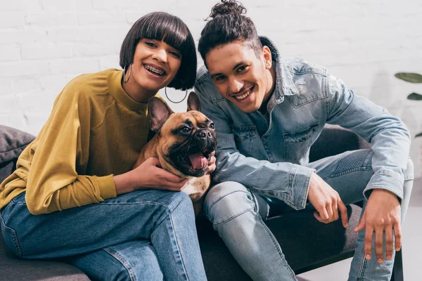 Smiling young mixed race couple sitting on couch with french bulldog — Stock Photo