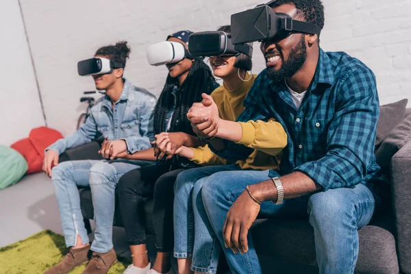 Young multicultural friends using virtual reality headsets and holding hands of each other — Stock Photo