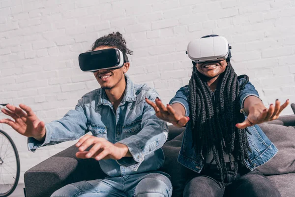 Молодые улыбающиеся друзья сидят на диване и используют гарнитуры виртуальной реальности — стоковое фото