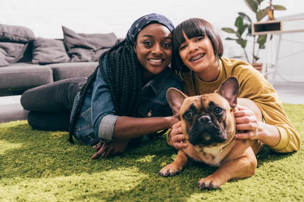 Two young multicultural female friends laying on rug with french bulldog — Stock Photo