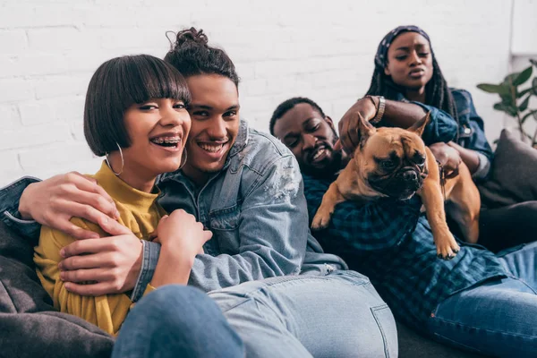 Joven grupo sonriente de amigos multiculturales sentados en el sofá con el perro - foto de stock