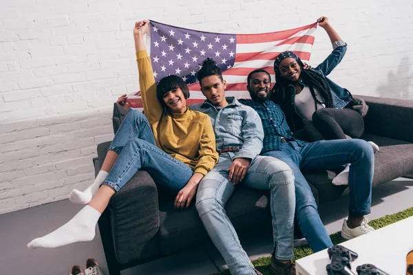 Group of young multicultural friends sitting on couch with flag of USA — Stock Photo