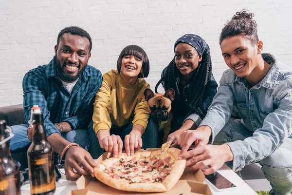 Группа улыбающихся молодых многонациональных друзей с французским бульдогом, принимающим пиццу — стоковое фото