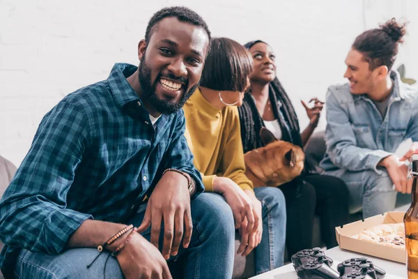 Souriant jeune homme noir assis sur le canapé avec des amis et un chien — Photo de stock