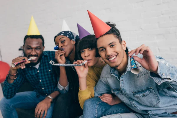 Young multiethnic friends in party hats celebrating with party horns — Stock Photo