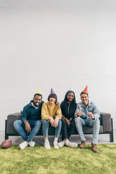 Группа улыбчивых молодых многонациональных друзей в шляпах, сидящих на диване — стоковое фото