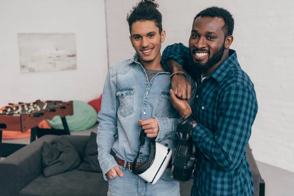 Two smiling multicultural young male friends standing with virtual reality headsets — Stock Photo