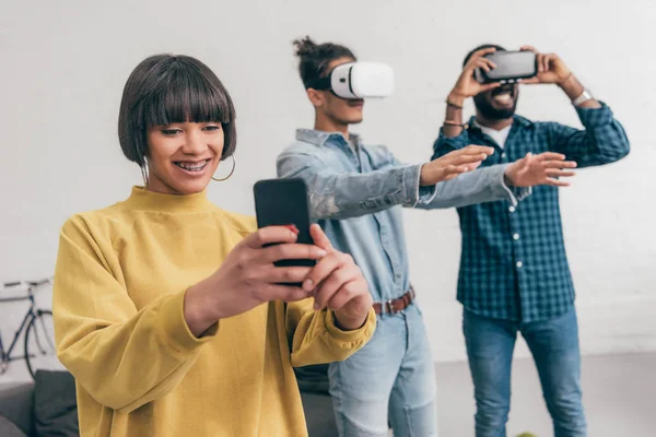 Souriant mixte jeune femme avec smartphone et deux hommes en utilisant des casques de réalité virtuelle derrière — Photo de stock