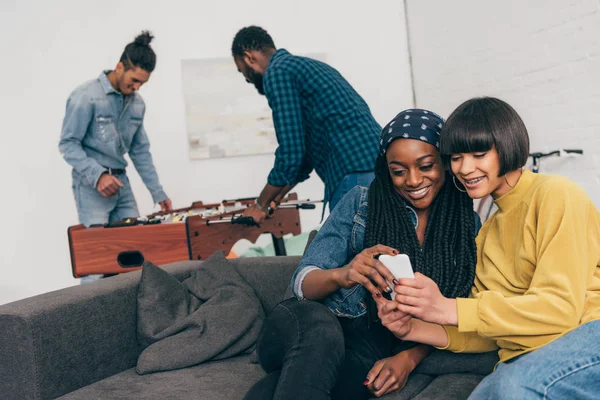 Two young female friends sitting with smartphone and two young men playing table soccer behind — Stock Photo