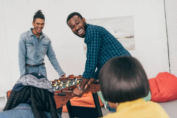 Laughing multiethnic friends playing table soccer and two female friends sitting beside — Stock Photo