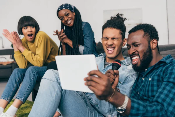 Zwei junge multiethnische männliche Freunde mit Mikrofon und digitalem Tablet singen Karaoke und Freundinnen sitzen dahinter — Stockfoto