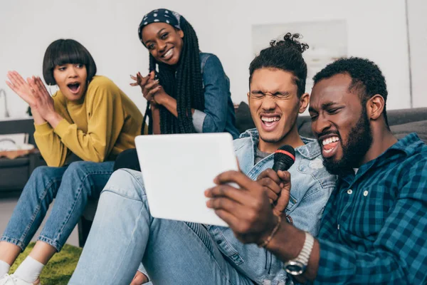 Dos jóvenes amigos masculinos multiétnicos con micrófono y tableta digital cantando karaoke y amigas sentadas detrás - foto de stock