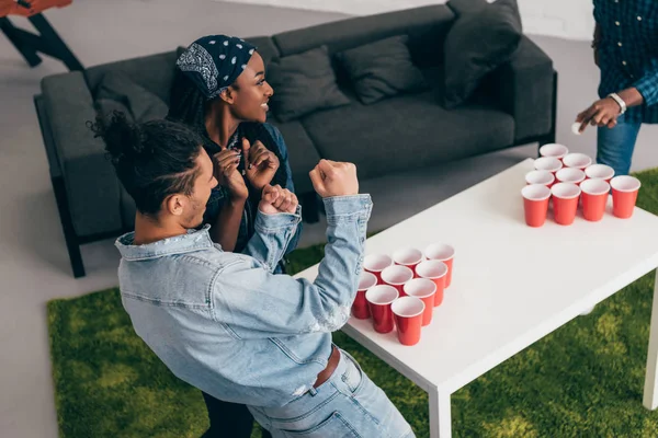 Recortado disparo de grupo multicultural de amigos jugando pong cerveza en la mesa y joven haciendo gesto ganador - foto de stock
