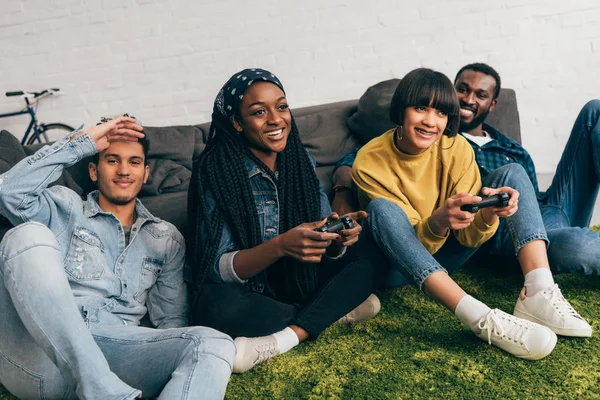 Dos mujeres jóvenes jugando videojuego por joysticks y amigos masculinos sentados al lado - foto de stock