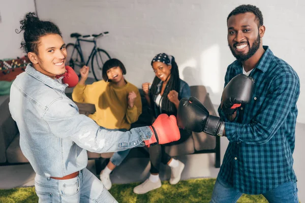 Smiling multicultural male friends doing sparring in boxing gloves and two women cheering up them — Stock Photo