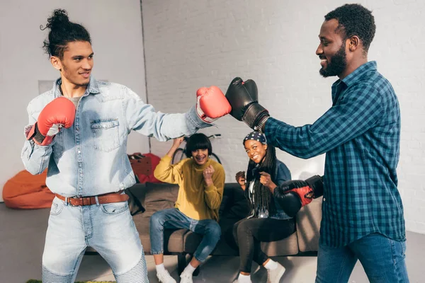 Multicultural male friends doing sparring in boxing gloves and two women cheering up them — Stock Photo