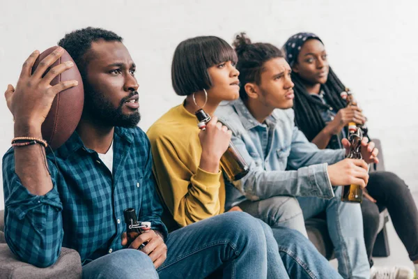 Засмучена мультикультурна група друзів сидить на дивані з пляшками пива і дивиться американський футбольний матч — стокове фото