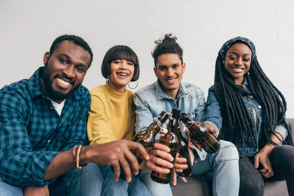 Quatre amis multiculturels souriants assis et cliquetis bouteilles de bière — Photo de stock