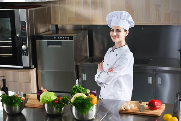 Chef feminino profissional de pé com braços dobrados por mesa com ingredientes culinários — Fotografia de Stock