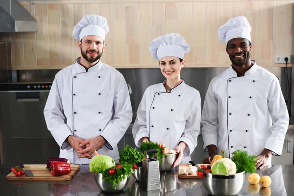 Equipo de chefs multirraciales sonriendo por el mostrador de cocina moderno - foto de stock