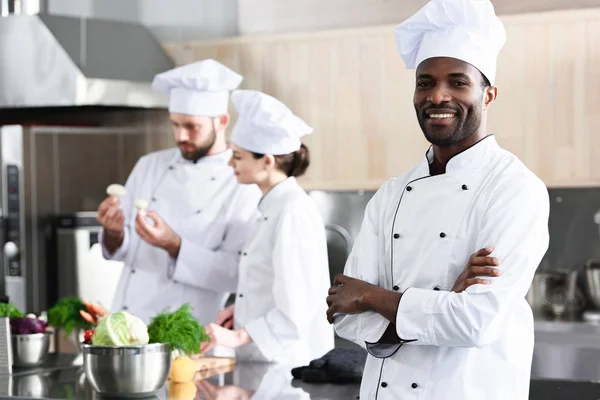 Chef afroamericano in piedi con le braccia incrociate di fronte ai suoi colleghi in cucina — Foto stock
